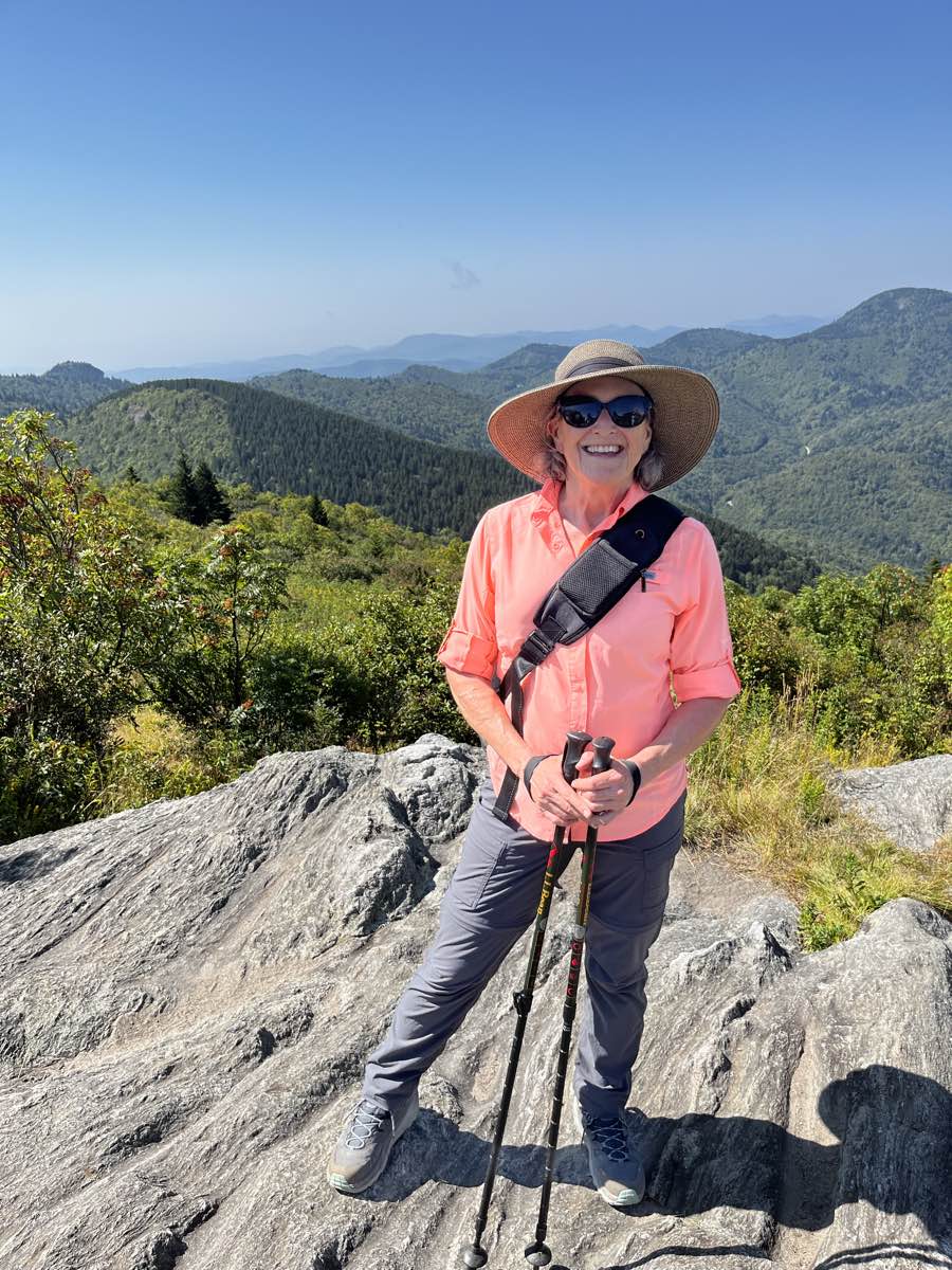 Sarah Cortvriend standing on a mountain hiking trail dressed in peach shirt and large hat and sunglasses.
