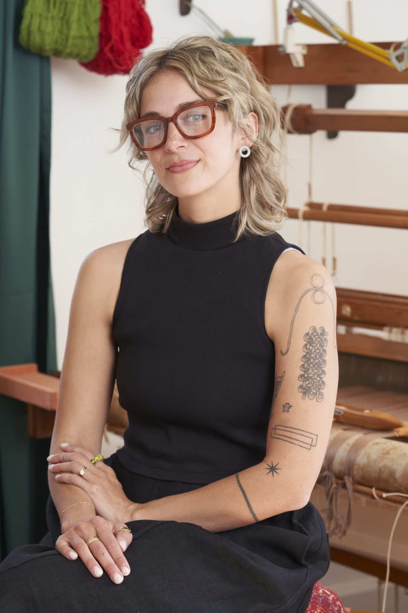 Woman with glasses and blonde hair sitting in front of a weaving loom, wearing black tank top, tattoos on arm.