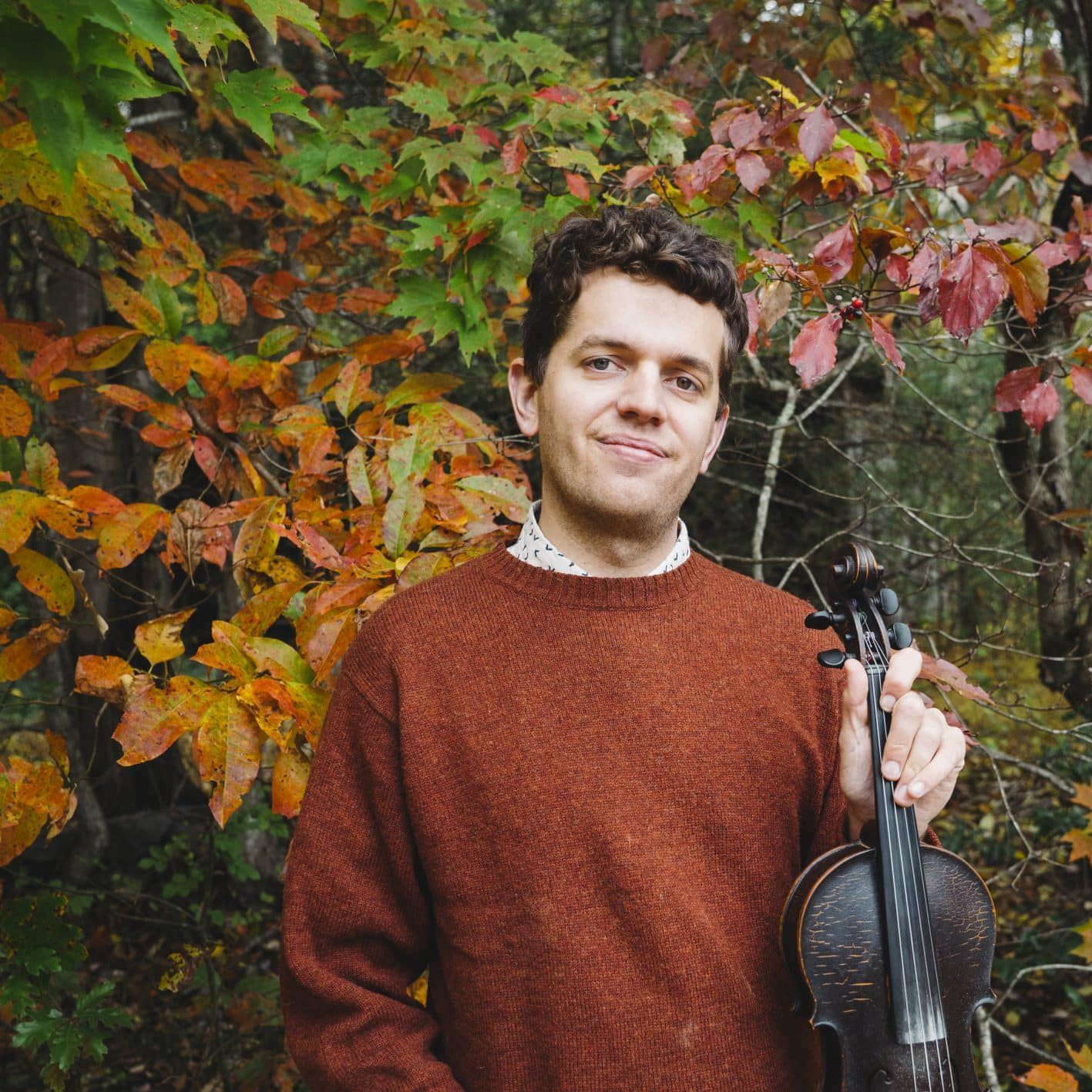 Image of a man holding a fiddle, dressed in a russet colored sweater with the changing leaves of autumn as a backdrop